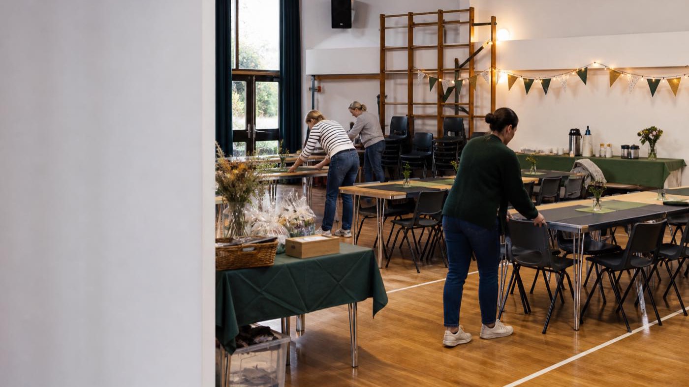 A school hall being set up for a summer fair, with folding tables, stacked chairs and bunting part way along the wall.